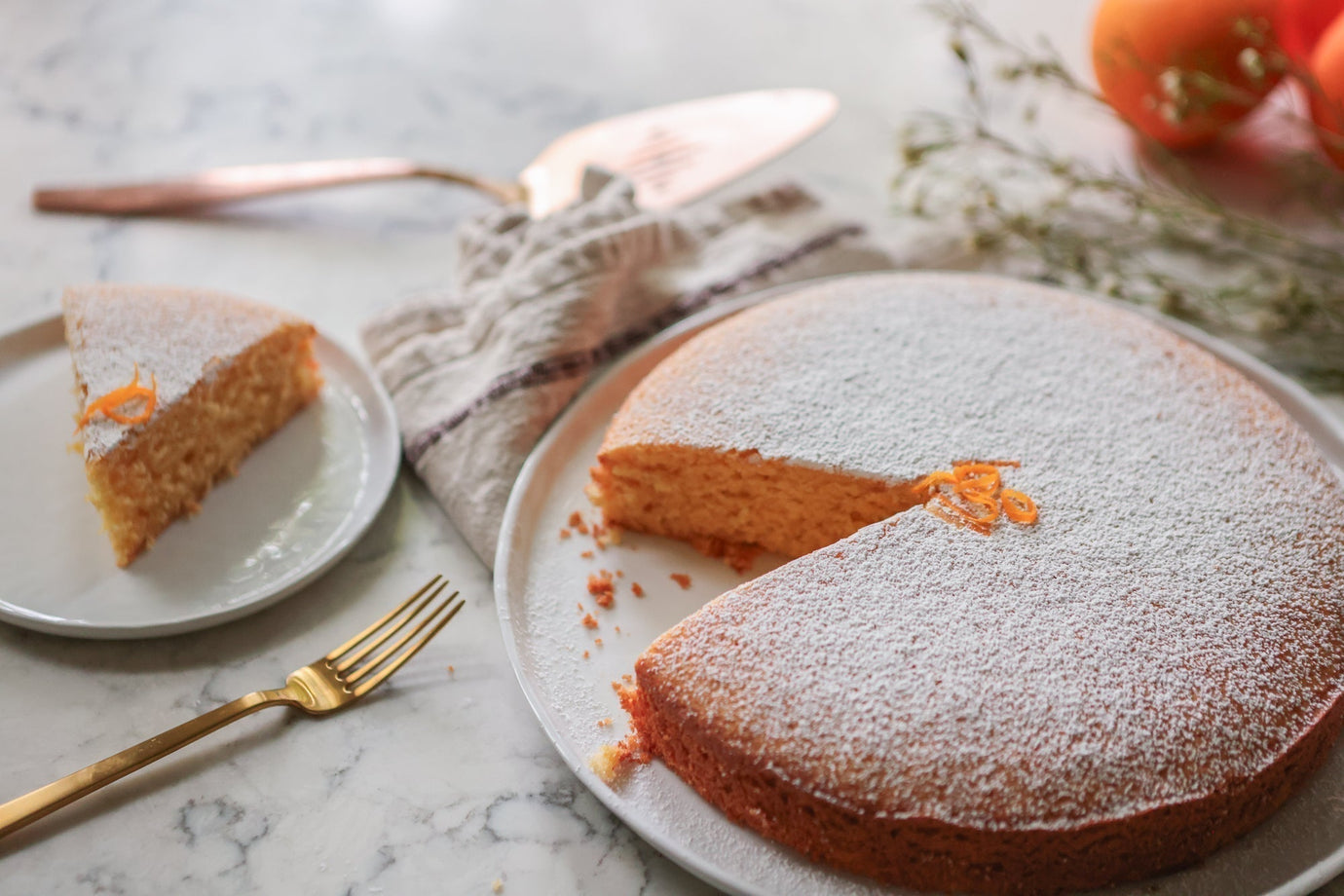 An image of a one layer orange cake with one slice cut from it on a plate with fresh oranges and flowers in the background